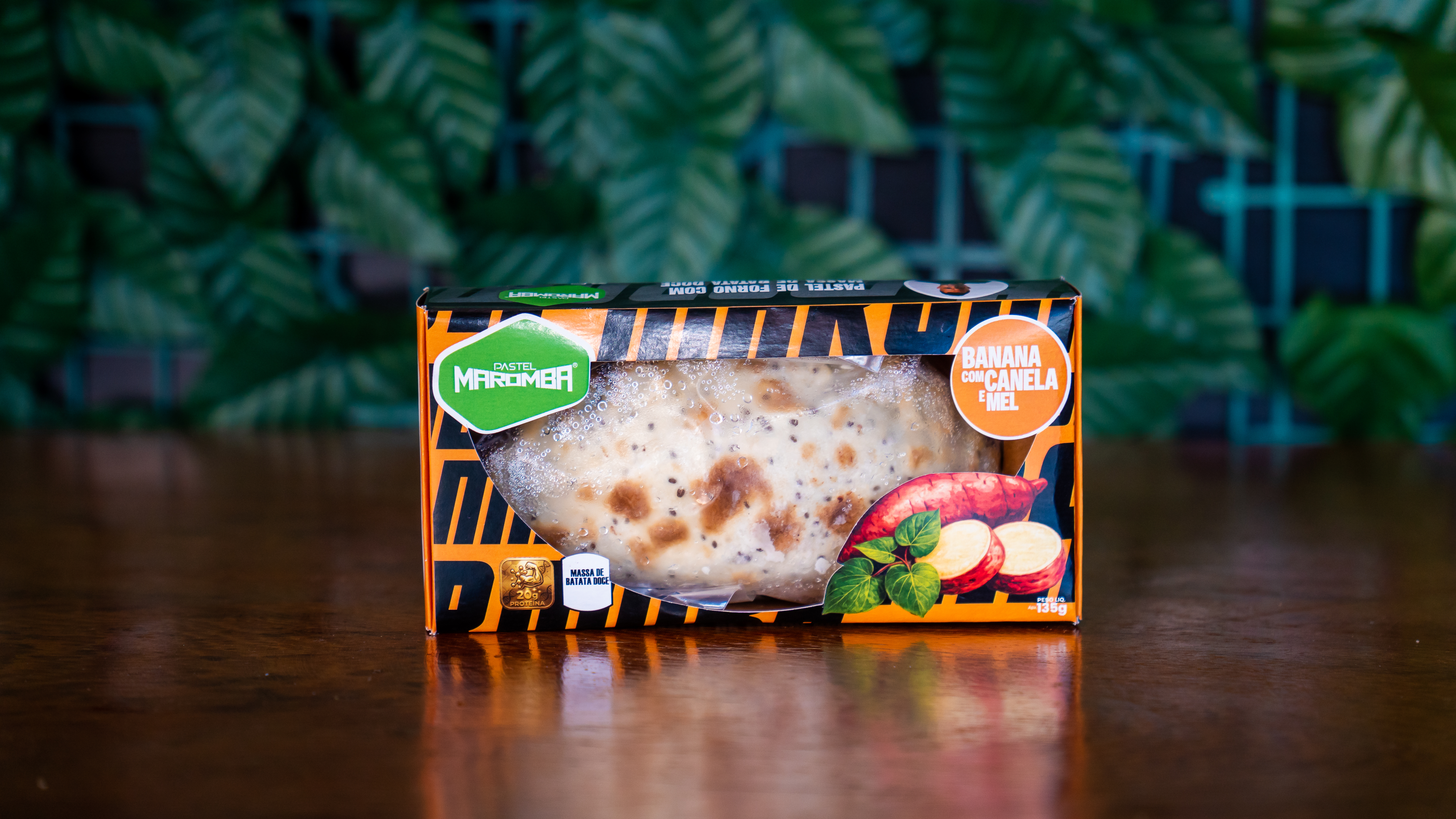 Caixa laranja de Pastel Maromba com selo Banana com Canela e Mel sobre mesa de madeira